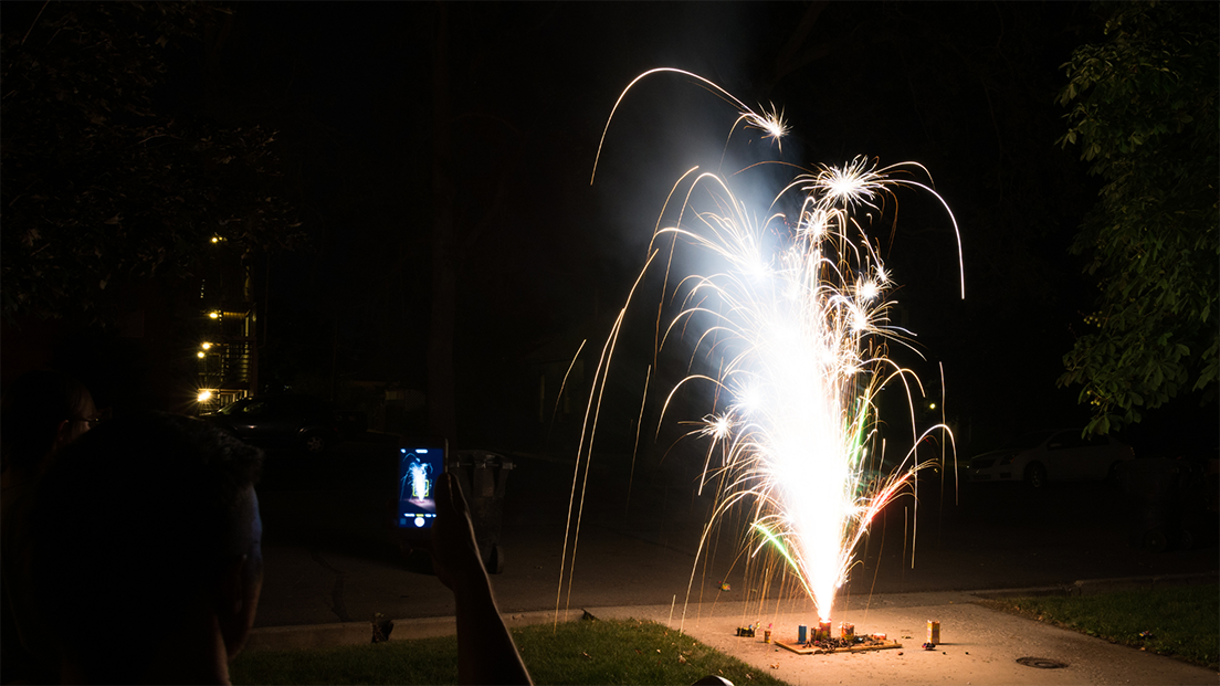 person holding phone while taking photo of small fireworks pyrotechnics display in driveway at night